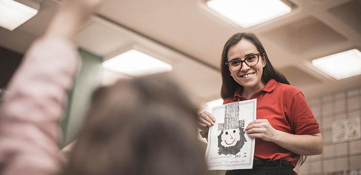 BSU student teacher smiling and holding up a childlike drawing of Abraham Lincoln while a student raises her hand